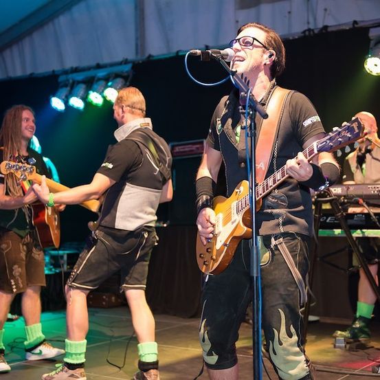 Eine Band tritt auf der Bühne auf, ein Gitarrist singt in ein Mikrofon, ein anderer Gitarrist spielt und ein Keyboarder im Hintergrund. Die Bühne ist mit farbigen Lichtern beleuchtet.