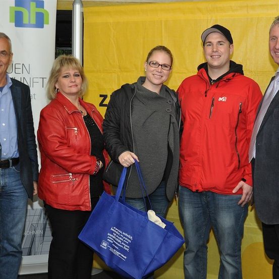 Fünf Personen stehen lächelnd vor einem gelben Hintergrund für ein Foto. Eine Frau in Rot hält eine blaue Tasche, während andere Jacken und Krawatten tragen.