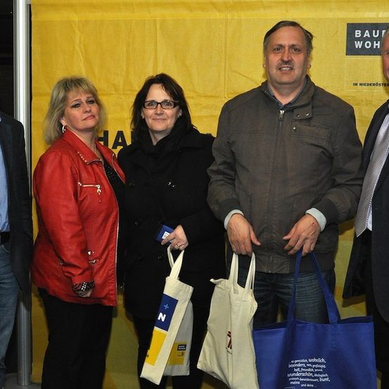 Fünf Personen stehen zusammen vor einem gelben Banner mit Text. Sie alle lächeln und halten Taschen. Der Mann rechts trägt eine Krawatte.
