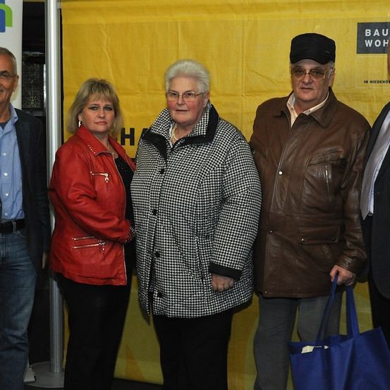 Fünf Personen stehen vor einem gelben Banner mit weißen Texten. Die Frau in der Mitte trägt einen schwarz-weiß karierten Mantel. Der Mann rechts hält eine blaue Tasche.
