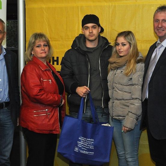 Eine Gruppe von Menschen, die für ein Foto zusammenstehen, zwei Frauen und zwei Männer, wobei einer einen blauen Beutel hält. Die Frau links trägt eine rote Jacke, die andere eine graue Jacke. Hinter ihnen ist ein gelber Banner.