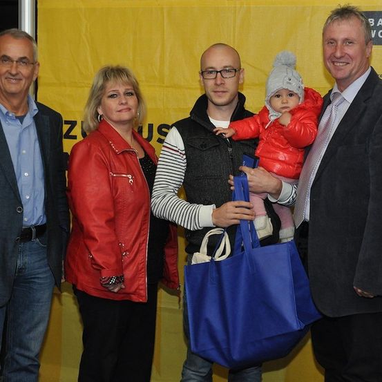 Fünf Menschen stehen vor einem gelben Banner. Ein Mann hält ein Baby in einem roten Mantel, während ein anderer Mann eine blaue Tasche hält. Die Frau im roten Mantel lächelt.
