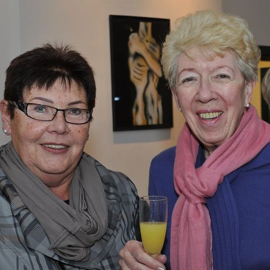 Zwei Frauen lächeln und halten ein Glas Orangensaft in einer Innenumgebung mit einem an der Wand montierten Kunstwerk im Hintergrund.