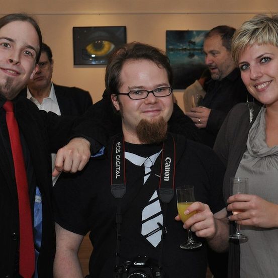 Drei Personen posieren für ein Foto bei einer Veranstaltung, ein Mann mit einer Kamera um den Hals, ein anderer mit einem Getränk und eine Frau lächelt.