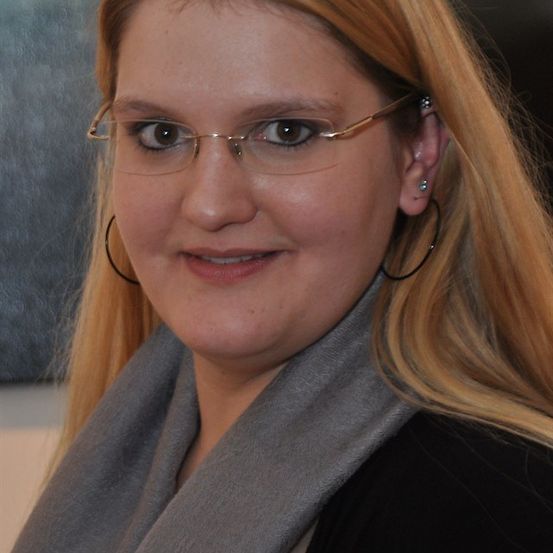 Eine Frau mit blonden Haaren, die eine Brille und einen grauen Schal trägt, lächelt und schaut nach vorne.