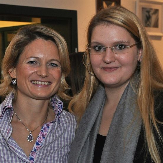 Zwei Frauen stehen eng beieinander und lächeln. Die Frau auf der linken Seite trägt ein Blumenhemd und eine Halskette. Die Frau auf der rechten Seite trägt eine Brille und einen grauen Schal.