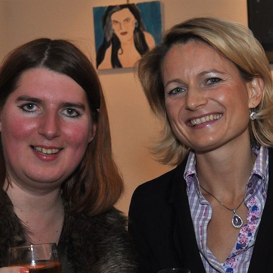 Zwei Frauen lächeln in die Kamera, eine hält ein Glas Wein in der Hand, vor einer Wand mit zwei Gemälden.