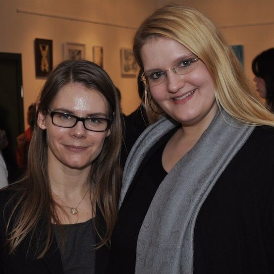 Zwei Frauen stehen eng beieinander, lächeln und posieren für ein Foto in einem Raum mit Kunst an den Wänden.
