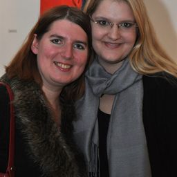 Zwei Frauen lächeln für ein Foto, eine mit Pelzkragen und die andere mit Brille und Schal.