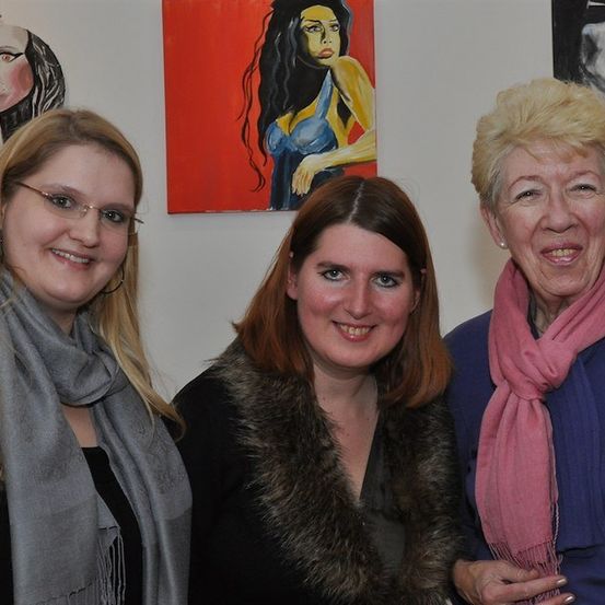 Drei Frauen stehen gemeinsam vor einer Wand mit Gemälden und lächeln für die Kamera. Die Frau auf der linken Seite trägt eine Brille und einen Schal, die Frau in der Mitte hat einen Pelzkragen und die Frau auf der rechten Seite trägt einen rosa Schal.