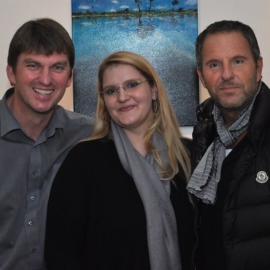 Drei Menschen stehen eng beieinander vor einem Gemälde und lächeln für ein Foto. Der Mann links trägt ein graues Hemd, die Frau in der Mitte hat eine Brille und einen Schal und der Mann rechts trägt einen schwarzen Mantel mit Logo.