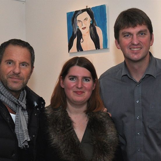Drei Menschen stehen vor einer Wand mit einem Gemälde einer Frau. Der Mann links trägt einen Schal und eine Jacke. Die Frau in der Mitte trägt einen Pelzmantel. Der Mann rechts trägt ein graues Hemd.