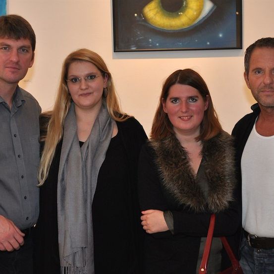 Vier Personen stehen zusammen vor einer Wand mit einem Gemälde eines gelben Auges. Der Mann links trägt ein graues Hemd, die Frau neben ihm trägt einen grauen Schal, die Frau rechts trägt eine mit Fell gefütterte Jacke und der Mann rechts trägt ein weißes Unterhemd.