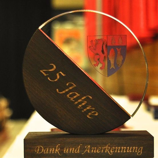 Eine hölzerne Trophäe mit Glasoberteil, eingraviert mit '25 Jahre' und einem Wappenschild, steht auf einem hölzernen Sockel mit den Worten 'Dank und Anerkennung'. Der Hintergrund ist mit roten Vorhängen verschwommen.