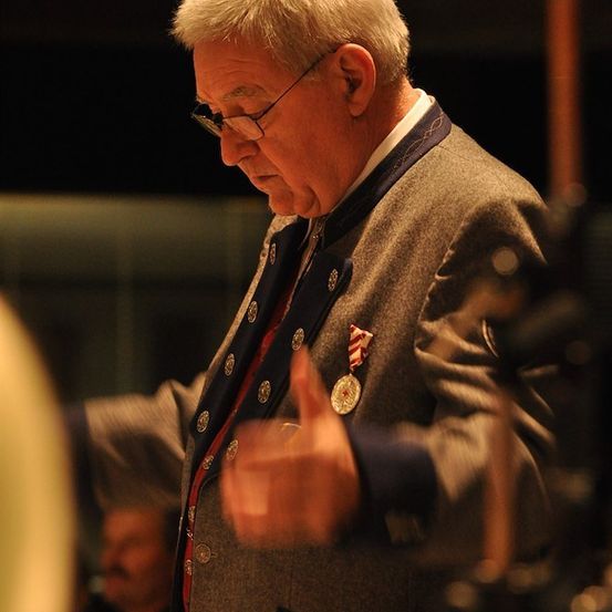 Ein älterer Mann mit Brille und Medaille dirigiert ein Orchester, seine Hand in der Luft erhoben.
