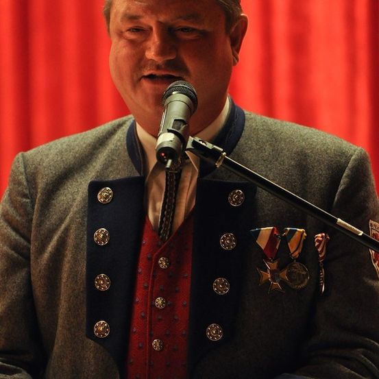 Ein Mann in einer dekorierten Uniform spricht in ein Mikrofon an einem Podium, mit einem roten Vorhang im Hintergrund.