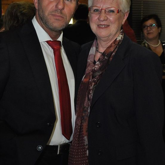 Ein Mann und eine Frau stehen eng beieinander und lächeln für ein Foto. Der Mann trägt eine rote Krawatte und die Frau hat ein gemustertes Tuch. Hinter ihnen schaut eine Frau mit Brille zu.