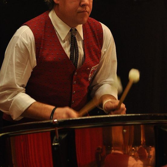 Ein Mann in einem roten Weste und weißem Hemd spielt eine Trommel mit einem Drumstick in der Hand.