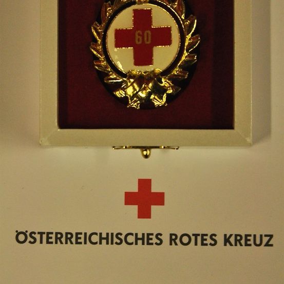 Eine gerahmte goldene Medaille mit einem roten Kreuz und der Nummer 60 darauf, auf einem weißen Hintergrund mit dem Text 'Osterreichisches Rotes Kreuz'.
