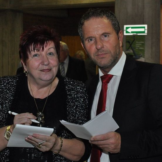 Ein Mann und eine Frau in formeller Kleidung stehen nebeneinander, halten Papiere und Stifte in der Hand, mit einem verschwommenen Hintergrund von Menschen und einem Schild.