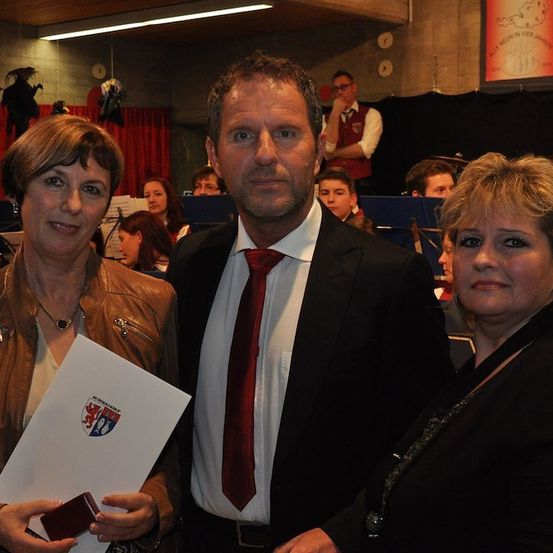 Ein Mann in Anzug und Krawatte steht zwischen zwei Frauen, hält ein Zertifikat und ein kleines Objekt. Im Hintergrund ist ein verschwommenes Publikum und eine Bühne zu sehen.