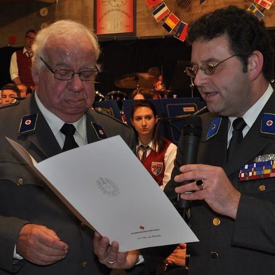 Zwei Männer in Militäruniformen, einer hält ein Papier und der andere ein Mikrofon, stehen vor einer Menschenmenge. Der Mann mit dem Mikrofon spricht.