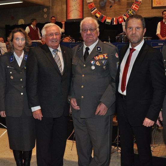 Fünf Personen in formeller Kleidung, einschließlich Militäruniformen, stehen zusammen in einem Raum mit Musikinstrumenten und Flaggen im Hintergrund.