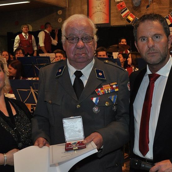 Ein Mann in Uniform hält eine Medaille und ein Zertifikat, flankiert von einer Frau und einem anderen Mann im Anzug. Dahinter spielt eine Band auf der Bühne.