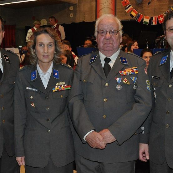 Vier Personen in Militäruniformen stehen zusammen, möglicherweise bei einer formellen Veranstaltung. Der Mann auf der rechten Seite trägt eine Brille und eine Medaille. Im Hintergrund sind verschwommen Personen und Flaggen zu sehen.