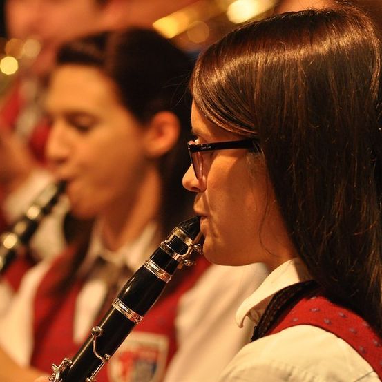 Zwei junge Frauen in roten Westen und mit Brille spielen Klarinetten in einer Band. Die Frau im Vordergrund konzentriert sich auf ihr Instrument.