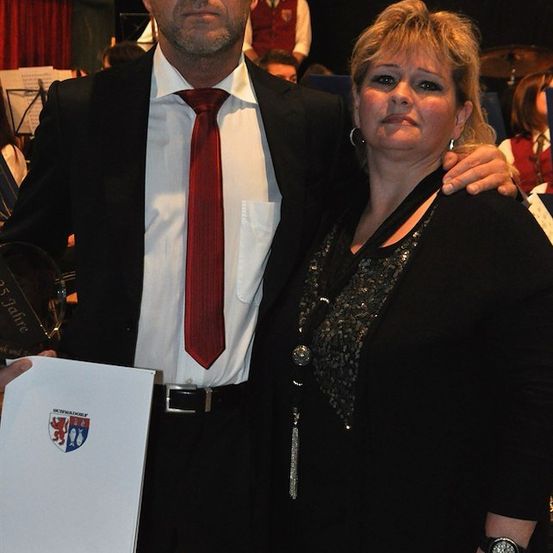 Ein Mann und eine Frau stehen zusammen, der Mann hält ein Zertifikat mit einem Logo, während die Frau in die Kamera schaut. Sie sind in einem Raum mit Musikinstrumenten und Leuten im Hintergrund.