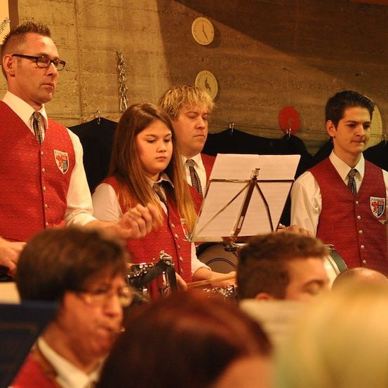 Eine Gruppe von Musikern, darunter ein Mann und eine Frau, spielt in einem Raum mit einer Betonwand und einer Uhr. Die Frau spielt die Trommel, während der Mann ein Notenpult hält.