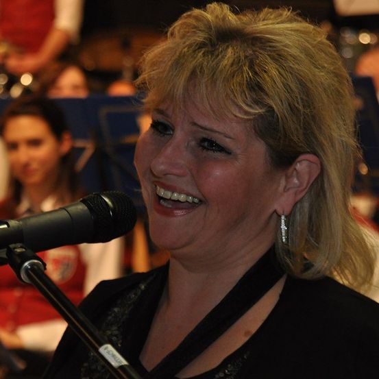 Eine Frau mit blonden Haaren lächelt und singt in ein Mikrofon. Sie trägt ein schwarzes Oberteil. Dahinter ist eine verschwommene Person mit einem roten Shirt zu sehen.