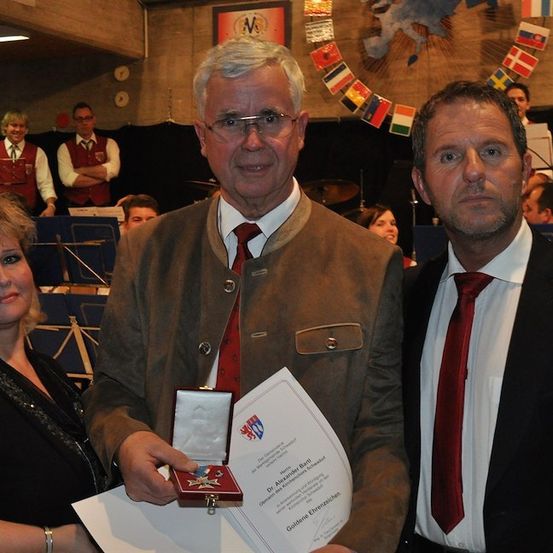 Ein Mann in einer braunen Jacke und roter Krawatte hält eine Medaille und ein Zertifikat, flankiert von einer Frau und einem anderen Mann in einem schwarzen Anzug und roter Krawatte, mit einer Band und Flaggen im Hintergrund.