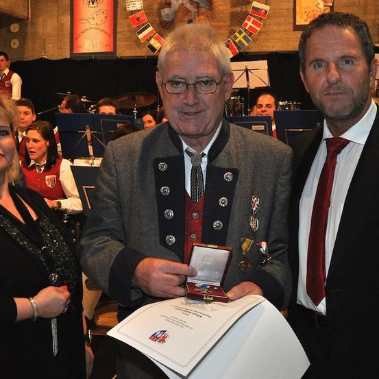 Ein Mann in traditioneller Uniform hält eine Auszeichnung und ein Zertifikat, flankiert von zwei anderen in formeller Kleidung, mit einer Band im Hintergrund.