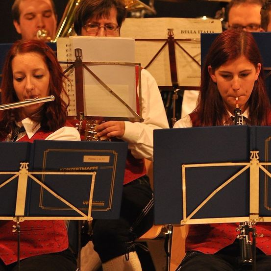 Eine Gruppe von Musikern spielt Instrumente bei einem Konzert. Eine Frau auf der linken Seite spielt Flöte und eine andere Frau auf der rechten Seite Klarinette. Sie tragen alle rote Westen. Hinter ihnen spielt ein Mann Posaune.
