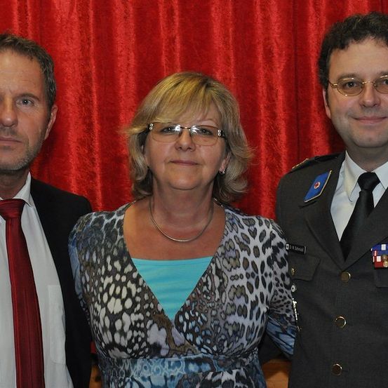Drei Personen stehen eng nebeneinander vor einem roten Vorhang. Der Mann links trägt eine rote Krawatte, die Frau in der Mitte trägt ein Leopardenmusterkleid und der Mann rechts trägt eine Militäruniform mit Medaillen.