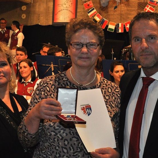 Eine Frau in einem Leopardenmusterkleid hält eine Medaille und ein Zertifikat, zwischen einem Mann in Anzug und einer Frau in einem schwarzen Kleid. Dahinter spielt eine Band auf der Bühne.
