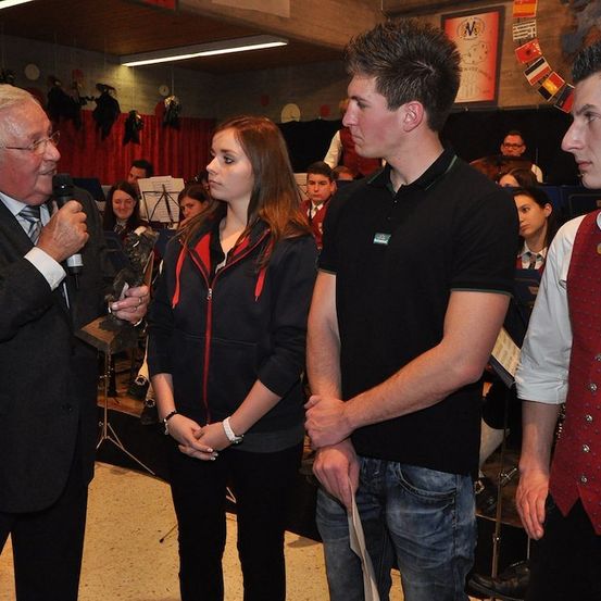 Ein älterer Mann mit Mikrofon spricht zu zwei jungen Leuten in einem Schulauditorium, mit einer Band und Publikum im Hintergrund.