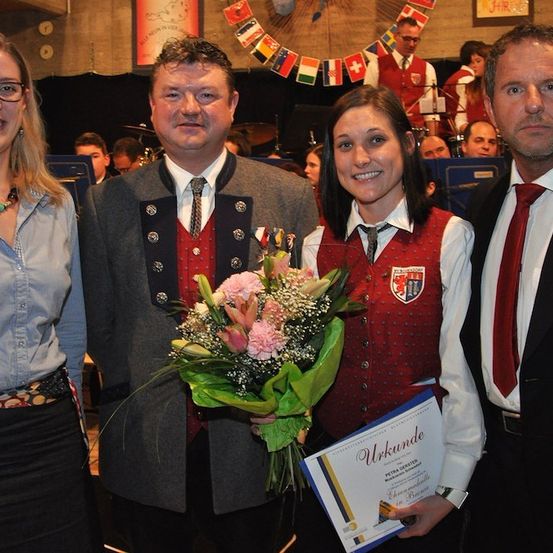 Eine Frau mit einem Blumenstrauß steht zwischen zwei Männern, einer hält ein Zertifikat, alle lächeln vor einem festlichen Hintergrund.