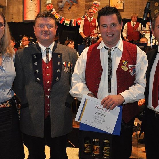 Vier Personen in traditioneller Kleidung stehen zusammen und lächeln für die Kamera. Der Mann in der Mitte hält ein Zertifikat und eine Weinbox. Hinter ihnen spielt eine Band, und Flaggen hängen an der Wand.
