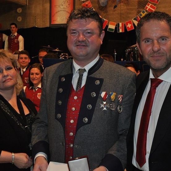 Drei Personen, zwei Männer und eine Frau, stehen bei einer Veranstaltung zusammen. Der Mann auf der linken Seite trägt ein traditionelles deutsches Outfit mit Medaillen. Der Mann auf der rechten Seite trägt einen Anzug und eine Krawatte. Hinter ihnen sind Menschen in roten Westen und Musikinstrumente zu sehen.