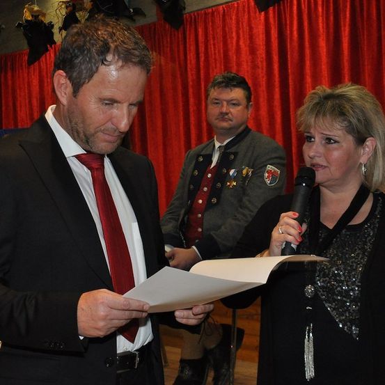Ein Mann in einem Anzug liest ein Blatt Papier, während eine Frau ein Mikrofon hält und ihn ansieht. Hinter ihnen steht ein weiterer Mann mit einem roten Band.