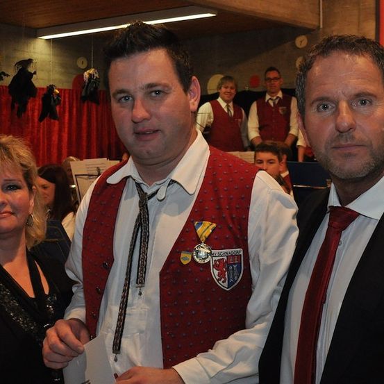 Drei Personen stehen in einem Raum zusammen. Der Mann in der Mitte trägt eine rote Weste mit Abzeichen und eine Krawatte. Der Mann rechts trägt einen schwarzen Anzug und eine rote Krawatte. Die Frau links trägt ein schwarzes Kleid. Hinter ihnen sitzen und stehen Leute.