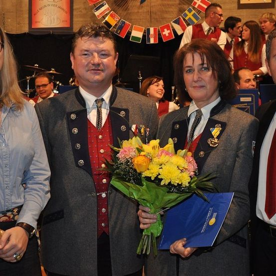 Eine Gruppe von vier Personen, zwei Männer und zwei Frauen, stehen zusammen und halten einen Blumenstrauß. Die beiden Männer tragen Anzüge und Krawatten, und die Frauen tragen Blazer. Sie alle lächeln und scheinen sich bei einer Veranstaltung zu befinden. Hinter ihnen stehen Leute und es hängen Flaggen an der Wand.