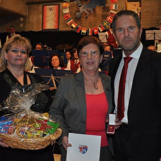 Drei Menschen stehen zusammen in einem Raum mit einer Bühne im Hintergrund. Die Frau auf der linken Seite hält einen Korb mit Snacks, die Frau in der Mitte hält ein Papier und der Mann auf der rechten Seite hält eine Schachtel. Hinter ihnen steht eine Bühne mit einer Band und einer Flaggendekoration.