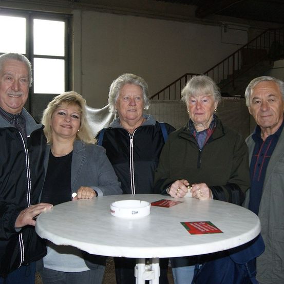 Fünf Menschen stehen um einen Tisch herum, lächeln und posieren für ein Foto. Auf dem Tisch stehen eine weiße Schüssel und rote Karten. Dahinter befindet sich eine Treppe und ein Fenster.