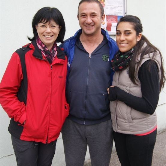 Drei Personen stehen zusammen vor einer weißen Wand und lächeln für ein Foto. Die Frau links trägt eine rote Jacke, der Mann in der Mitte eine blaue Jacke und die Frau rechts eine beige Weste.