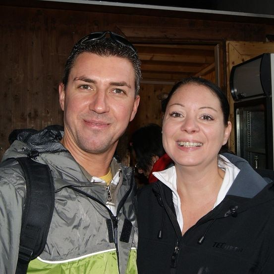 Ein Mann und eine Frau lächeln und stehen eng beieinander, möglicherweise für ein Foto. Der Mann trägt einen Rucksack und eine Jacke. Hinter ihnen ist eine Holzwand und ein Kühlschrank.