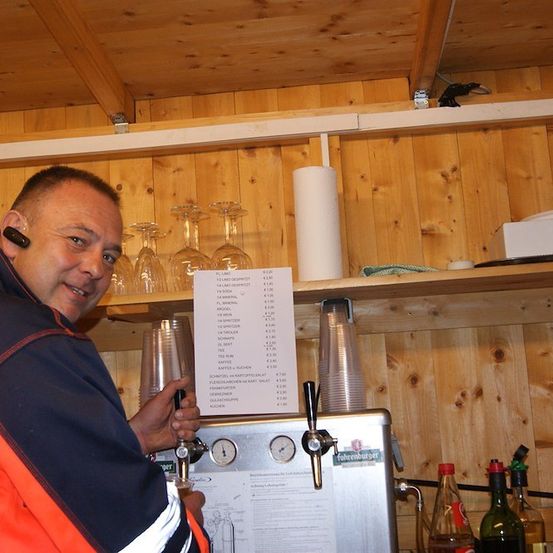 Ein Mann in einer blau-orangefarbenen Jacke lächelt und hält einen Bierhahn in einer hölzernen Bar. Ein Menü befindet sich über ihm auf einem Regal.